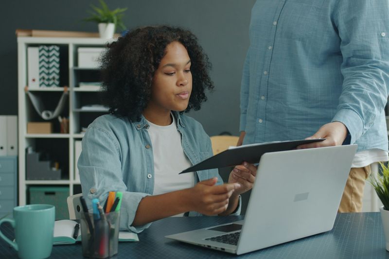 A woman at a laptop looks through a document while a second person hands her another page.