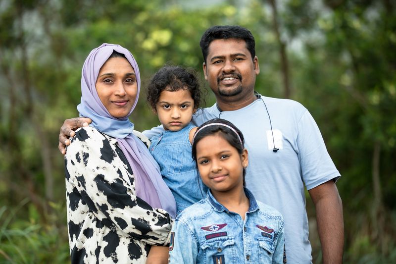 A mother in a headscarf, a father and their two young daughters stand together outdoors.