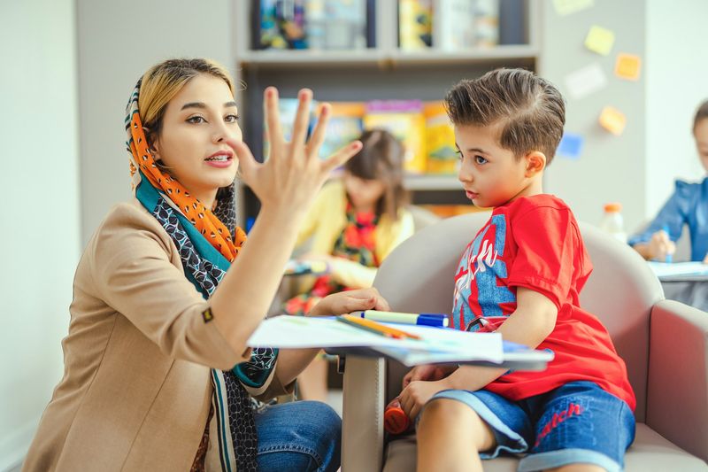 A woman in a headscarf explains something with her hands to an attentive young boy at a table.