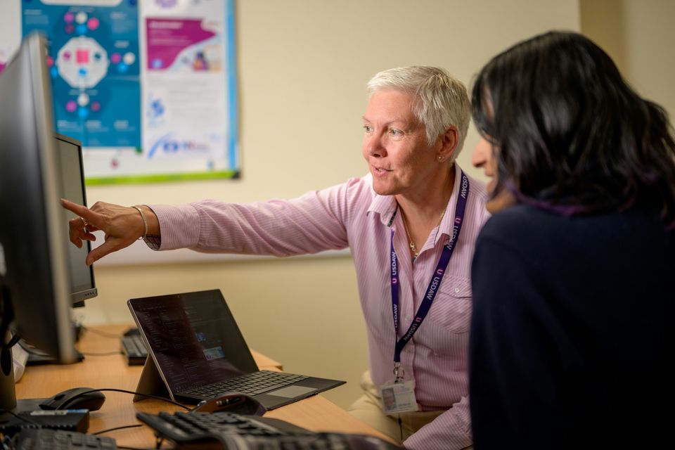 A woman sits beside a visitor at a desk and points to information on a computer screen.
