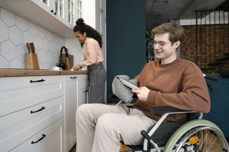 A man in a wheelchair reads in his kitchen while a woman prepares food in the background.