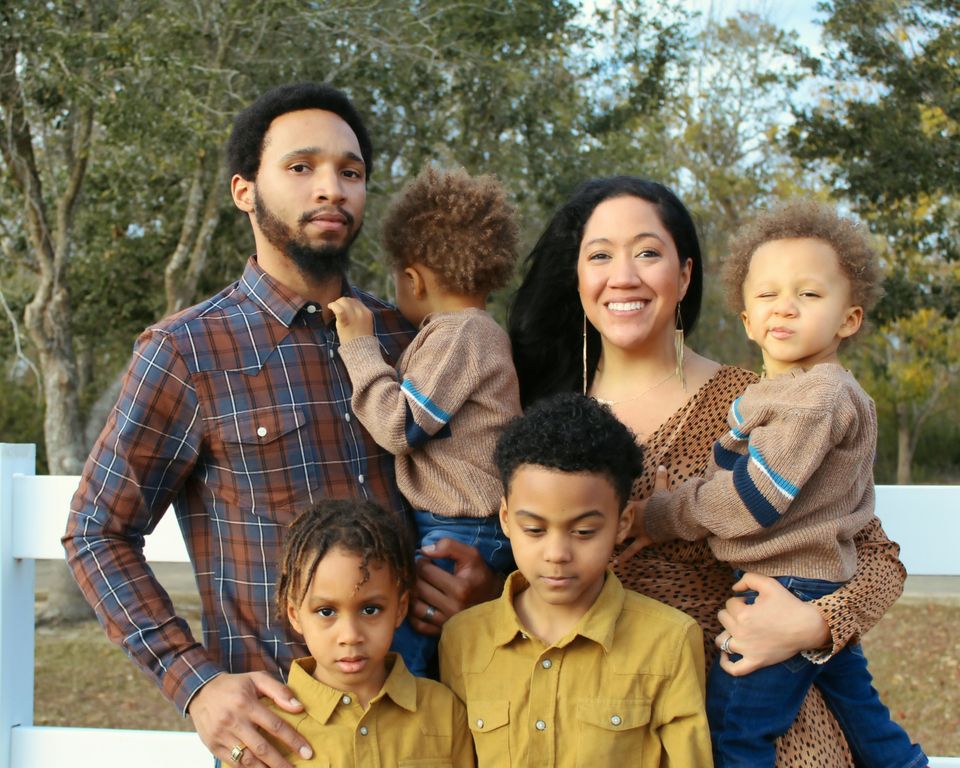 A father, mother and four young children stand close together outdoors, smiling in warm afternoon light.