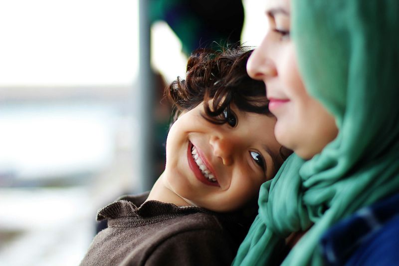 A smiling boy leans his head against his mother, who wears a green headscarf.