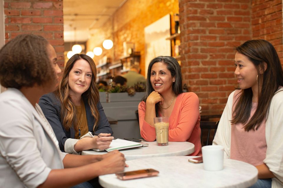 Four women talk around a small café table with notebooks and coffee.