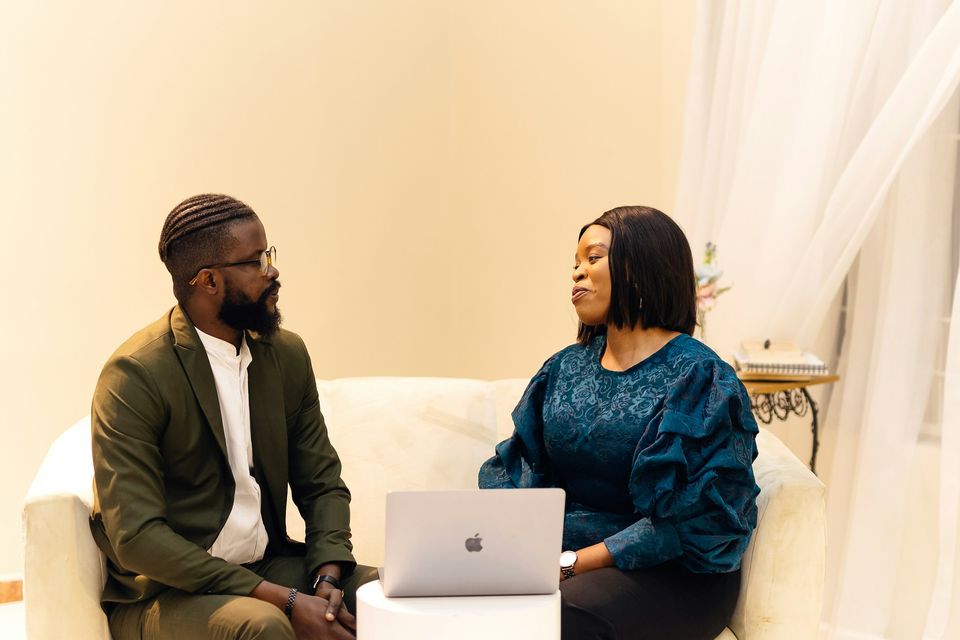 A man and a woman in business clothes sit facing each other in conversation, a laptop between them.