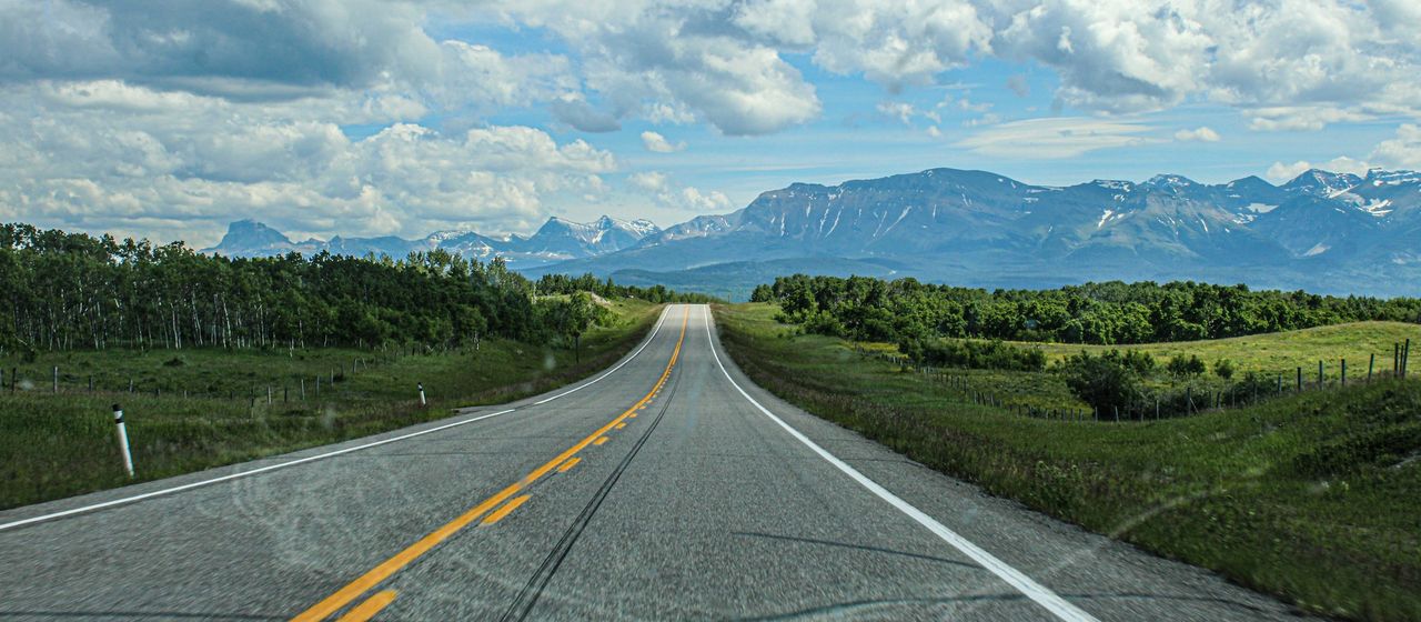 A two-lane highway runs straight across green foothills toward the Rocky Mountains.