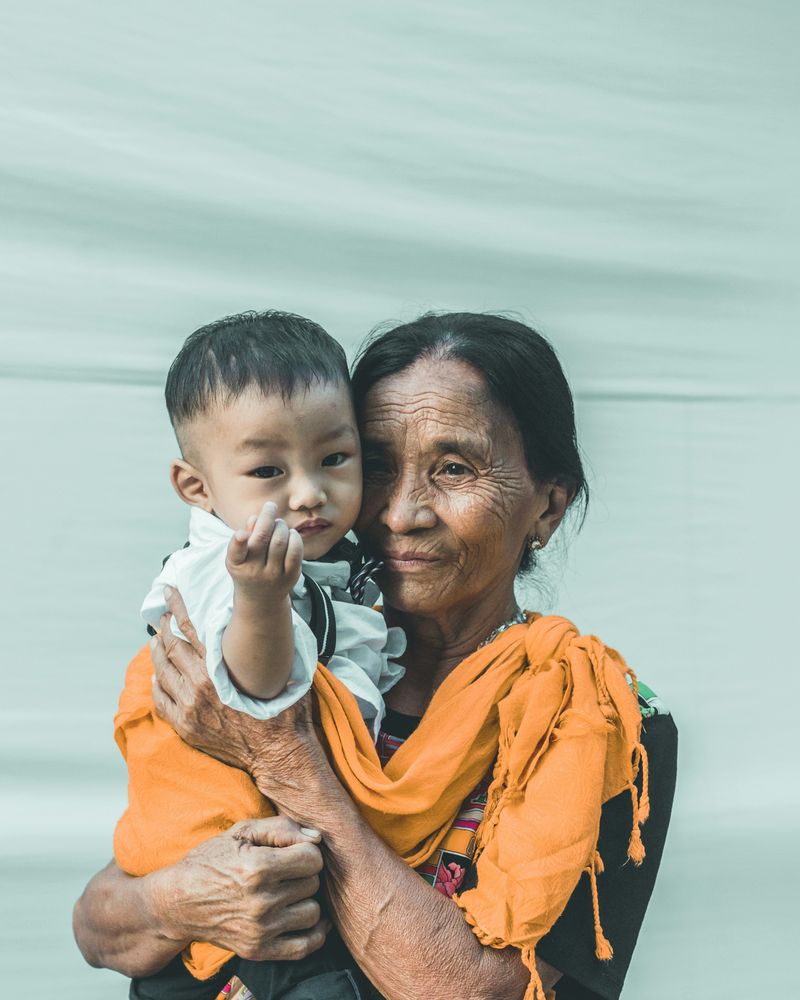 An older woman in a bright orange scarf holds a toddler on her hip.