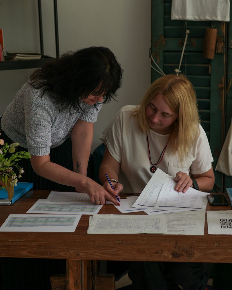 Two women sit side by side at a wooden table, reviewing printed documents together.