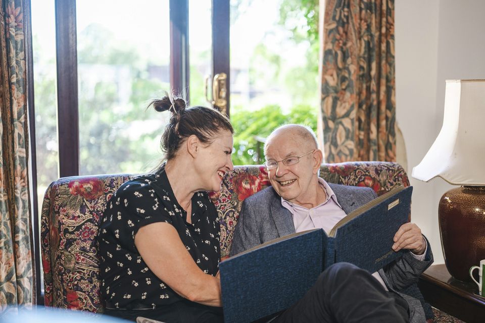 A woman and an older man laugh together while looking through a photo album on a sofa.