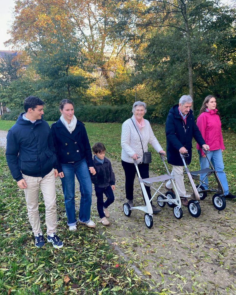 Three generations of a family walk together on an autumn path; one grandparent uses a walker.